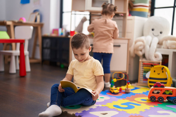Adorable toddler reading book sitting on floor at kindergarten - Fotó, kép