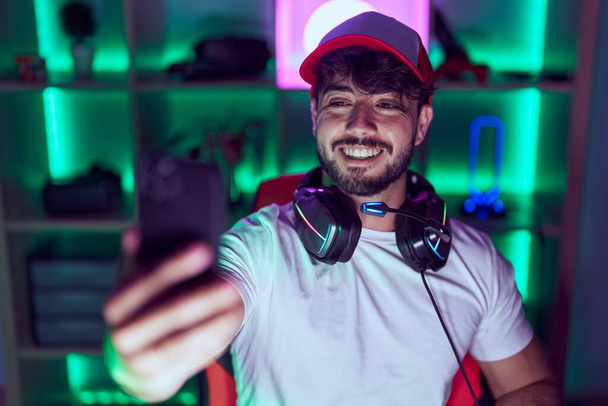 Joven hombre hispano streamer usando computadora hacer selfie por smarpthone en gamin habitación - Foto, imagen