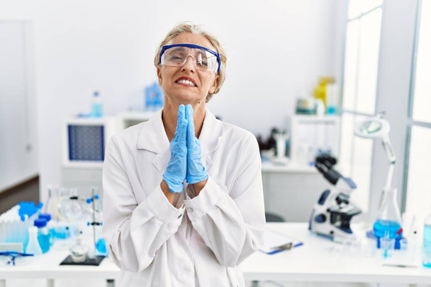 Keski-ikäinen blondi nainen työskentelee tiedemies laboratoriossa kerjäämässä ja rukoilemassa kädet yhdessä toivon ilme kasvoilla hyvin emotionaalinen ja huolissaan. kerjääminen.  - Valokuva, kuva