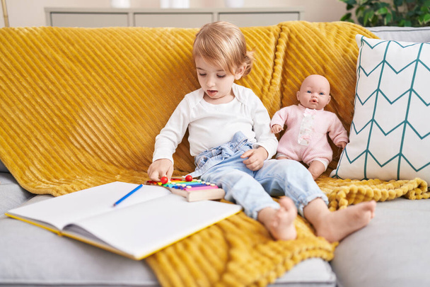 Adorable caucasian girl playing xylophone sitting on sofa with baby doll at home - Foto, imagen