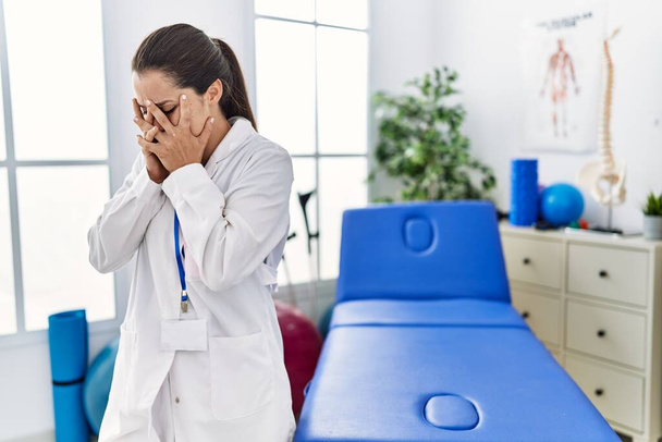 Jonge doktersvrouw die werkt in een pijnkliniek met een droevige uitdrukking en gezicht bedekt met handen terwijl ze huilt. depressie concept.  - Foto, afbeelding