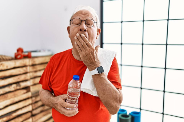 Hombre mayor con ropa deportiva y toalla en el gimnasio aburrido bostezo cansado cubriendo la boca con la mano. inquietud y somnolencia.  - Foto, imagen