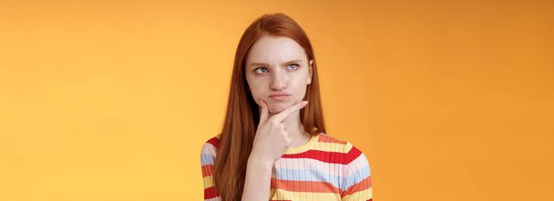 Hämmentynyt epäröivä vakavan näköinen inkivääri tyttö siniset silmät vieroitus valintoja pouting touch leuka näyttää vasemmassa yläkulmassa harkittuja ajattelu ymmällään ratkaisu, pohtiminen päätös, oranssi tausta. - Valokuva, kuva