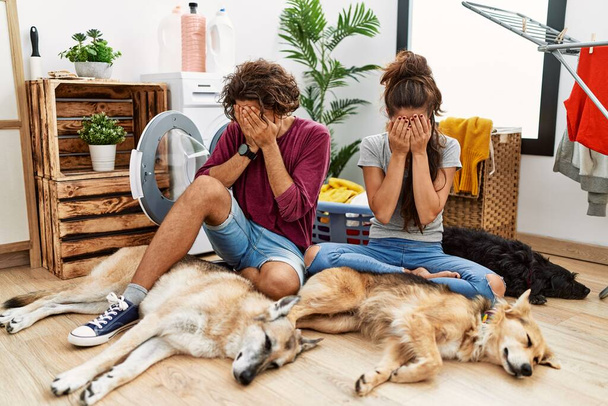 Νεαρό ζευγάρι ισπανόφωνων κάνει μπουγάδα με σκυλιά με λυπημένη έκφραση που καλύπτει το πρόσωπο με τα χέρια, ενώ κλαίει. έννοια της κατάθλιψης.  - Φωτογραφία, εικόνα