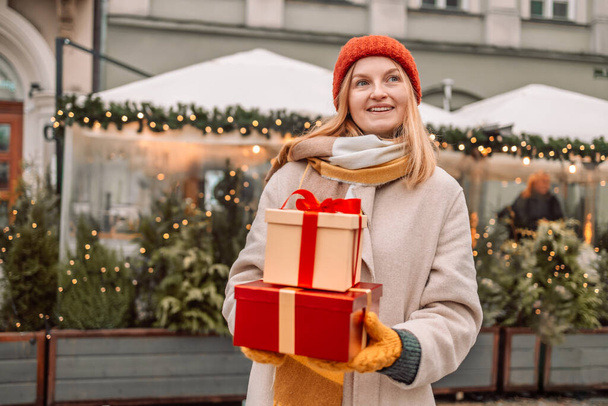 Счастливая молодая привлекательная женщина с бумажными пакетами и подарочными коробками в руках после Рождества покупки в торговом центре. Веселая красивая девушка держит сумки для покупок и представляет коробки в руках. Зимние каникулы - Фото, изображение