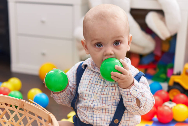 Adorable bebé caucásico chupando pelota sentado en el suelo en el jardín de infantes - Foto, Imagen