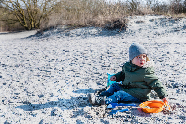 Hevesli hevesli bir çocuk kumsalda oturup kışın kum oyuncaklarıyla oynuyor. Ateşli ya da ateşli çocuk soğuk havaya karşı giyinmiş ya da örtülmüş bir iplikte oturuyor, güneşli bir kış gününde oynuyor. - Fotoğraf, Görsel