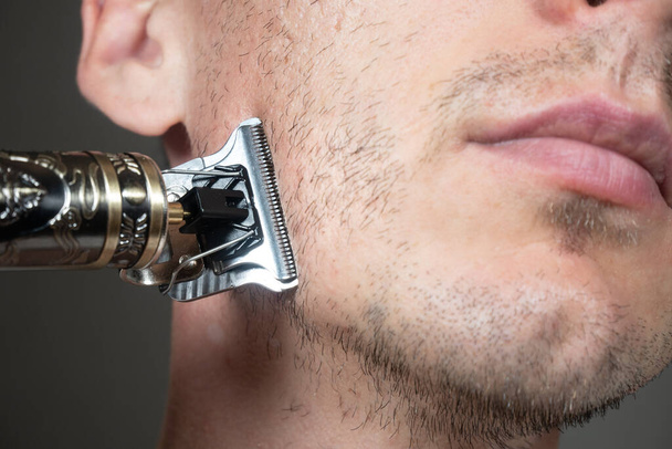 De mens snijdt baard door Elektrisch metalen scheermes of droog scheerapparaat. Trimmer voor snijden. Korte, dunne baard op het gezicht van een man. Haargroeiproblemen. Ongeschoren haren op de baard. - Foto, afbeelding