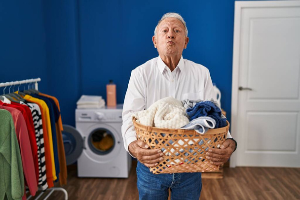Ein älterer Mann mit grauen Haaren, der zu Hause Wäschekorb hält und in die Kamera blickt, bläst einen Kuss, der schön und sexy ist. Liebesausdruck.  - Foto, Bild