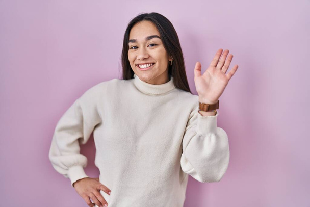 Young south asian woman standing over pink background waiving saying hello happy and smiling, friendly welcome gesture  - Photo, Image