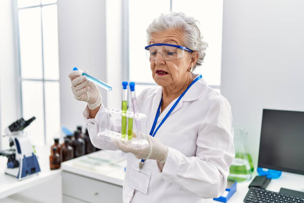 Idősebb ősz hajú nő, tudós egyenruhát visel, kémcsöveket tart a laboratóriumban. - Fotó, kép
