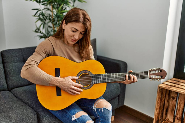 Middle age caucasian woman playing classical guitar sitting on the sofa at home. - Photo, Image