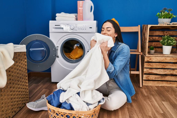 Jovem bela mulher hispânica sorrindo confiante cheirando roupas limpas na lavanderia - Foto, Imagem
