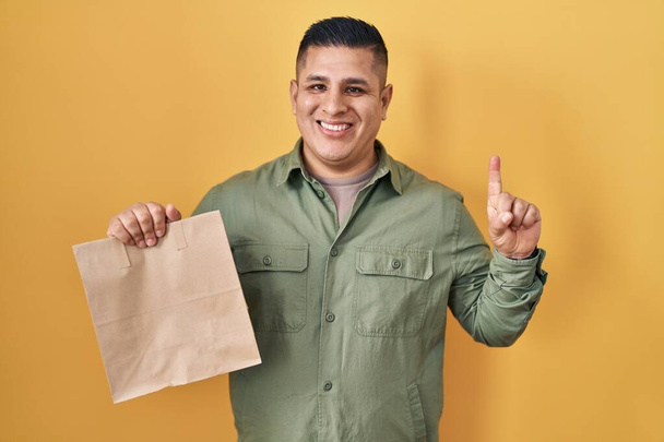 Latino nuori mies tilalla ottaa pois paperipussi hymyillen idea tai kysymys sormella onnellinen kasvot, numero yksi  - Valokuva, kuva