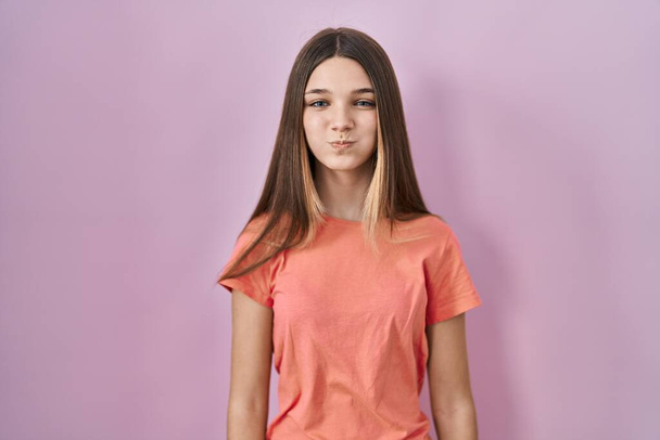 Teenager κορίτσι στέκεται πάνω από ροζ φόντο puffing μάγουλα με αστείο πρόσωπο. στόμα φουσκωμένο με αέρα, τρελή έκφραση.  - Φωτογραφία, εικόνα