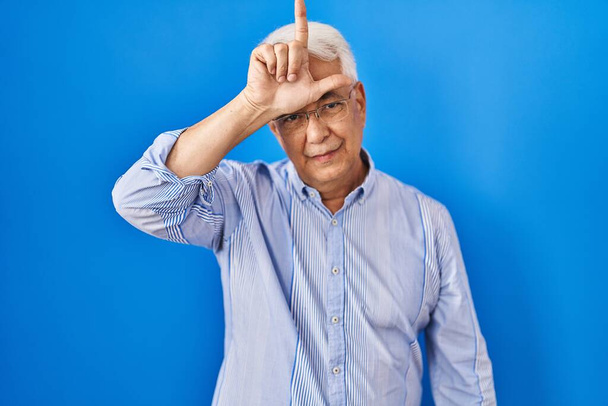 Hispanique homme âgé portant des lunettes se moquant des gens avec les doigts sur le front faisant geste perdant moquerie et insultant.  - Photo, image