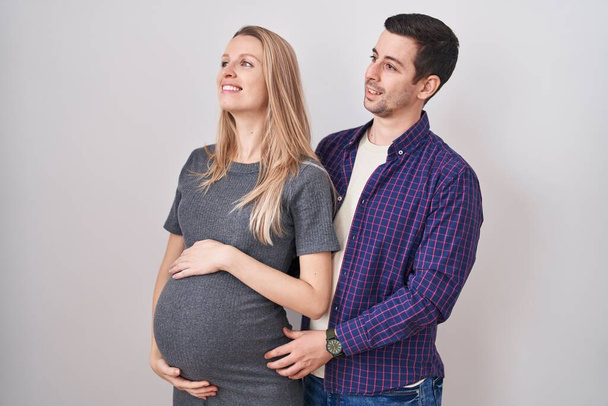 Junges Paar, das ein Baby erwartet, das vor weißem Hintergrund steht und mit einem Lächeln im Gesicht und natürlichem Ausdruck zur Seite schaut. Lachen selbstbewusst.  - Foto, Bild