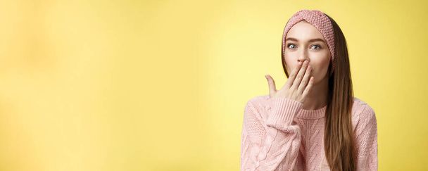 Geschwätzige junge nette Büroangestellte im Pullover, Strickstirnband mit Handfläche auf dem Mund überrascht und erstaunt, erfährt interessante schockierende Gerüchte, Klatsch und Tratsch unterhalten über die gelbe Wand. - Foto, Bild