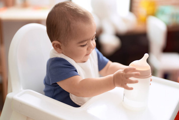 Entzückendes chinesisches Kleinkind mit Fütterungsflasche sitzt auf Essensstuhl Baby zu Hause - Foto, Bild