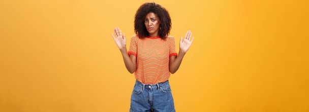 不安と悲しいです黒肌の女性モデルで流行の縞模様のTシャツとショートパンツで腕を上げて降伏眉をひそめます関与していないとオレンジ色の背景に無関心に立って - 写真・画像