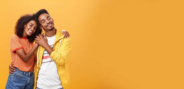 Ze houden van elkaar. Portret van twee charmante Afrikaans-Amerikaanse man en vrouw in relatie knuffelend met hartverwarmende glimlach aandoenlijke handen lachend zachtjes poserend tegen oranje achtergrond. - Foto, afbeelding
