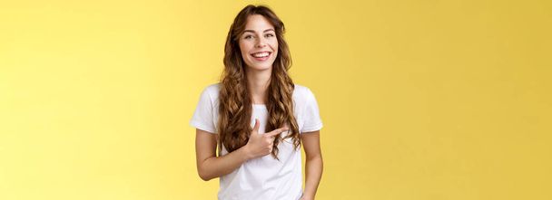 Έξοδος γοητευτικό όμορφη γυναίκα σγουρά μακρύ χτένισμα δείχνοντας δείκτη αριστερά δείχνουν διαφήμιση γέλιο ξένοιαστα ικανοποιημένοι κρατήστε το χέρι τσέπη casual φιλικό ποζάρουν κίτρινο φόντο. - Φωτογραφία, εικόνα