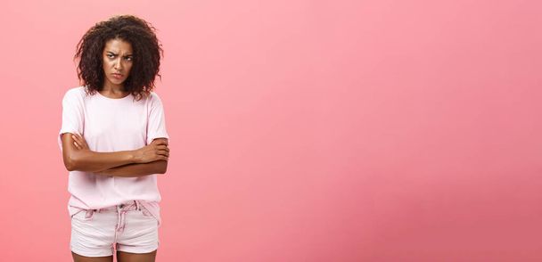 Porträt einer gekränkten, düster traurigen afrikanisch-amerikanischen Freundin, die die Arme auf der Brust verschränkt, in schützender Geste die Stirn runzelt und beleidigt über die rosa Wand blickt - Foto, Bild