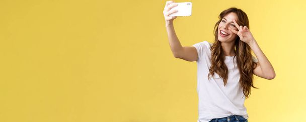 Radosny charyzmatyczny przystojny kręcone włosy kobieta biały t-shirt pokazać pokój zwycięstwo znak uśmiechnięty szeroko stwarzające przetargu przyjazny wyraz smartfon przedni aparat podjąć selfie żółty tło - Zdjęcie, obraz