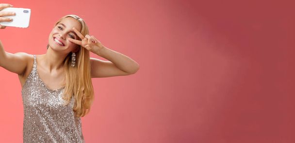Styl życia. Stylowy wspaniały urok młoda blondynka w błyszczącej srebrnej sukience przechyla głowę beztroski pokaz pokój gest zwycięstwo znak przedłużyć ramię trzymając smartfon biorąc selfie nagrywania wideo post - Zdjęcie, obraz