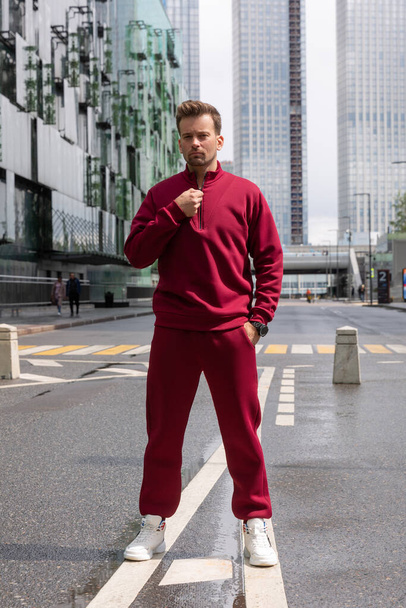 Full-length young man wears a vinous sports suit and poses for the camera. Athletic handsome young man stands on urban location and shows sports suit. Mock-up - Photo, image