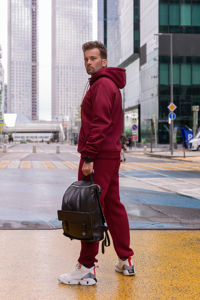 Full-length young man wears a vinous red sports suit and poses for the camera, he holds a backpack in his hands. Athletic handsome young man stands on urban location and shows sports suit. Mock-up - Фото, изображение