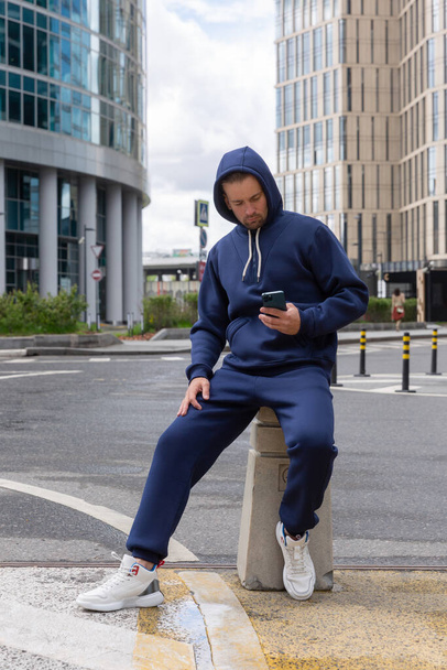 Full-length young man wears a blue sports suit and Looks at the phone. Athletic handsome young man sits on urban location and shows sports suit. Mock-up - Foto, Imagem