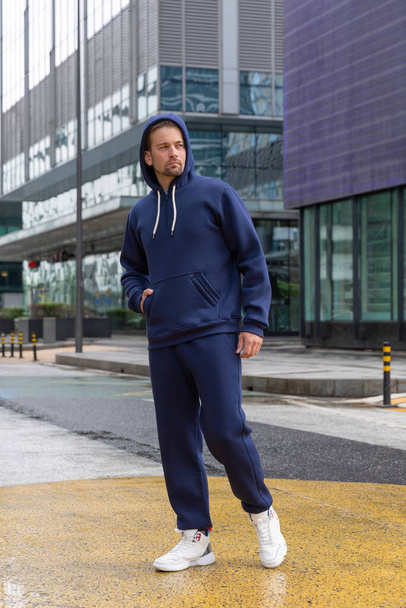 Full-length young man wears a blue sports suit and poses for the camera. Athletic handsome young man stands on urban location and shows sports suit. Mock-up - Fotoğraf, Görsel