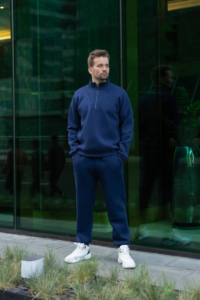 Full-length young man wears a vinous sports suit and poses for the camera. Athletic handsome young man stands on urban location and shows sports suit. Mock-up - Foto, Imagem