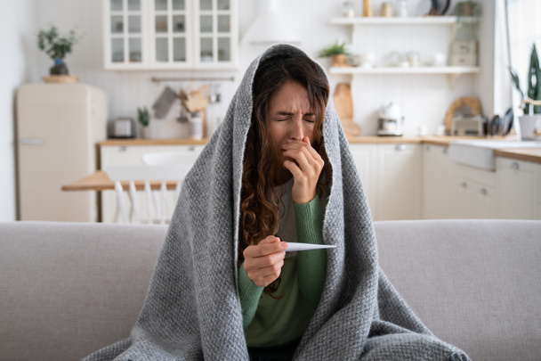 鼻水、くしゃみに苦しんで自宅でソファに座っている女性。毛布に包まれた病気の女性は、温度を測定する温度計を保持し、風邪やインフルエンザの症状の影響を受けています。冬のウイルス - 写真・画像