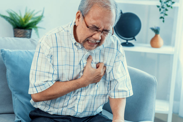An agonizing senior man suffering from chest pain or heart attack alone in his living room. Serious health problem and feeling unwell concept for seniors. - Foto, Imagem