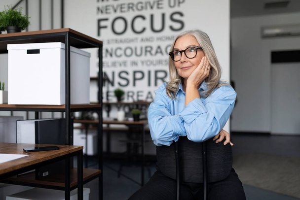 mujer de negocios mayor caucásica en una camisa azul y gafas se sienta pensando en la oficina. - Foto, imagen