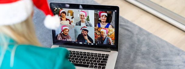 Улыбающаяся девушка в шляпе Санты, которая обзванивает семью веб-камерой. Женщина с ноутбуком сидит на кухне дома, используя виртуальный чат встреч по праздникам. С Новым годом и Рождеством! - Фото, изображение