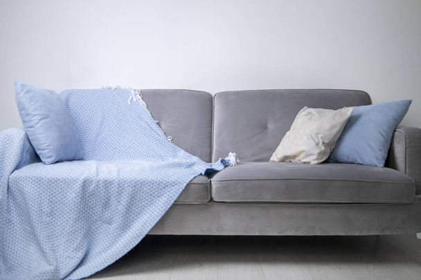Em um sofá cinza encontra-se uma colcha azul e travesseiros decorativos contra uma parede branca. - Foto, Imagem