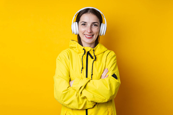 Uśmiechnięta młoda kobieta w słuchawkach ze skrzyżowanymi ramionami w żółtej kurtce na żółtym tle - Zdjęcie, obraz