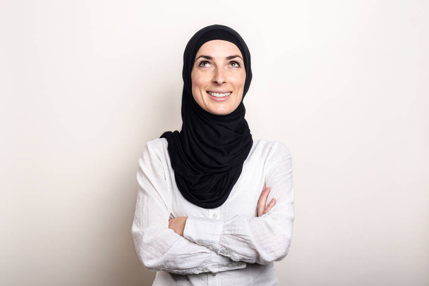 Young Muslim woman dressed in a white shirt and hijab looks up on a light background. Banner. - Photo, Image