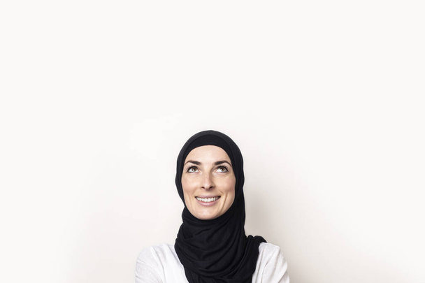 Νεαρή Μουσουλμάνα ντυμένη με λευκό πουκάμισο και χιτζάμπ κοιτάζει πάνω σε ελαφρύ φόντο. Μπάνερ. - Φωτογραφία, εικόνα