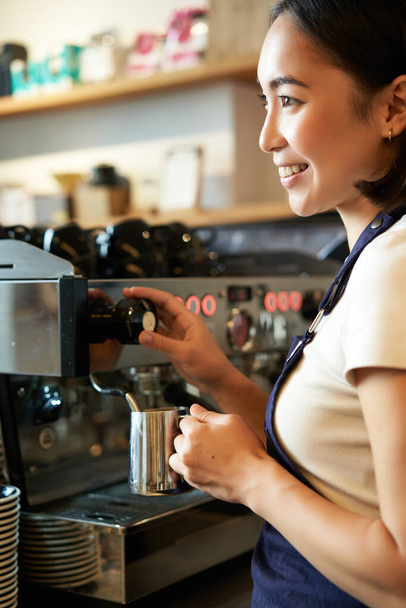 Щаслива азіатка, бариста використовує кавоварку для замовлення, готує молоко для капучино і латте, сміється і посміхається під час роботи в кафе
. - Фото, зображення
