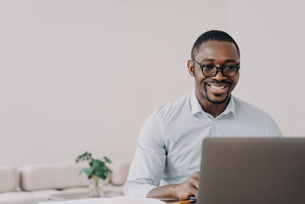 アフリカ系アメリカ人の男性ソフトウェア開発者がラップトップでオンラインでプロジェクトに取り組んで眼鏡をかけて笑っています。幸せな黒の男のプログラマーは良いニュースで現代的なアプリケーションや読書メールを作成します. - 写真・画像