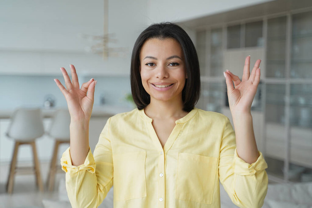 Smiling young woman keeping calm with mudra gesture, practicing yoga breathing exercises at home. Serene female relaxing her mind for stress relief. Healthy lifestyle, wellness, emotional control. - Photo, Image