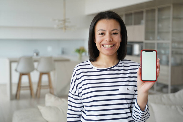 Ευτυχισμένη γυναίκα δείχνει smartphone με κενή οθόνη mock up, στέκεται στο σπίτι. Σύγχρονη χαμογελώντας θηλυκό κρατώντας τηλέφωνο με άδεια οθόνη πρότυπο, συνιστά νέες εφαρμογές, μεγάλες εκπτώσεις στο ηλεκτρονικό κατάστημα. - Φωτογραφία, εικόνα