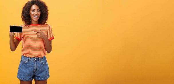 Egy lány, aki a telefonban mutogatja a menő nyaralását. Bájos barátságos és társaságkedvelő afro-amerikai nő göndör haj gazdaság mobiltelefon mutatva okostelefon képernyő felett narancssárga fal - Fotó, kép