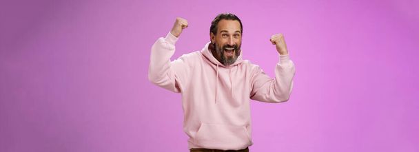 Yeah who cares age. Carefree delighted happy old man bearded in trendy pink hoodie raise fists joyfully triumphing having fun glad win celebrating success accomplish goal, posing purple background. - Photo, Image