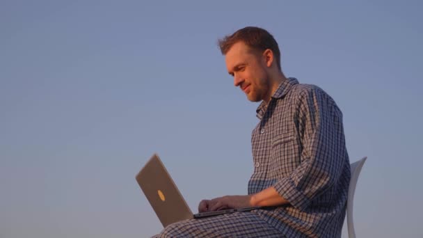 Trabajando en concepto matutino. Adorable barbudo caucásico freelancer masculino que trabaja al aire libre usando el ordenador portátil con pijamas escribiendo y luego mirando hacia adelante, pensando. Imágenes de alta calidad 4k - Imágenes, Vídeo