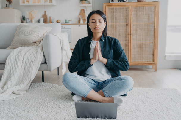 オンラインヨガのレッスン中にノートパソコンで瞑想する女性は、自宅で床に蓮の花に座っている。穏やかな女性の音声の肯定を聞く静かさを感じる。健康、落ち着き、ストレス軽減の概念. - 写真・画像